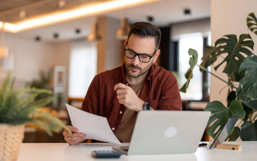 Man in an office environment with a silver laptop looking at a piece of white paper